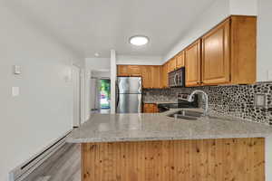 Kitchen featuring a baseboard heating unit, decorative backsplash, light stone countertops, appliances with stainless steel finishes, and brown cabinetry