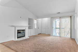 Unfurnished living room featuring vaulted ceiling, a glass covered fireplace, built in features, and light colored carpet