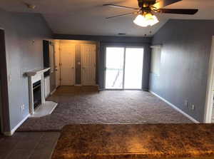 Unfurnished living room with vaulted ceiling, ceiling fan, dark carpet, dark tile patterned flooring, and a glass covered fireplace