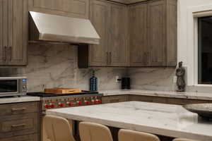 Kitchen with under cabinet range hood, dark brown cabinetry, appliances with stainless steel finishes, light stone countertops, and decorative backsplash