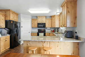 Kitchen with light brown cabinets, black appliances, a peninsula, light stone counters, and a textured ceiling