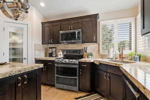 Kitchen featuring dark brown cabinets, stainless steel appliances, light stone countertops, backsplash, and light wood finished floors
