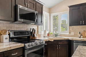 Kitchen with appliances with stainless steel finishes, tasteful backsplash, dark brown cabinetry, lofted ceiling, and light stone countertops