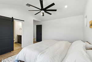Bedroom featuring a barn door, lofted ceiling, light wood-type flooring, a ceiling fan, and ensuite bath