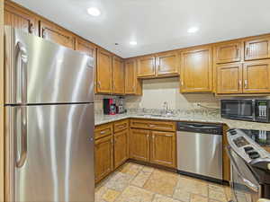 Kitchen featuring stainless steel appliances, brown cabinets, recessed lighting, stone tile floors, and light stone counters