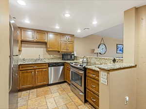 Kitchen with brown cabinetry, a peninsula, stainless steel appliances, light stone counters, and recessed lighting