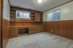 Unfurnished office featuring wood walls, wainscoting, built in desk, crown molding, and light colored carpet