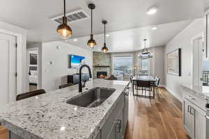 Kitchen with gray cabinetry, a fireplace, dark wood-style flooring, hanging light fixtures, and a center island with sink