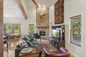 Living area with light wood-style flooring, a stone fireplace, a towering ceiling, healthy amount of natural light, and beam ceiling
