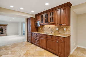Kitchen featuring decorative backsplash, brown cabinetry, light stone countertops, glass insert cabinets, and a brick fireplace