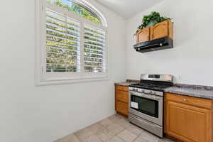 Kitchen with stainless steel gas range, under cabinet range hood, brown cabinets, and dark countertops