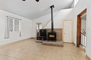 Unfurnished living room with a wood stove, vaulted ceiling, healthy amount of natural light, light tile patterned floors, and ceiling fan