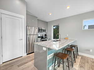 Kitchen with a breakfast bar area, gray cabinets, appliances with stainless steel finishes, light wood finished floors, and light stone counters