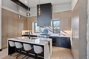 Kitchen with dark cabinets, light wood finished floors, pendant lighting, light stone counters, and a center island with sink