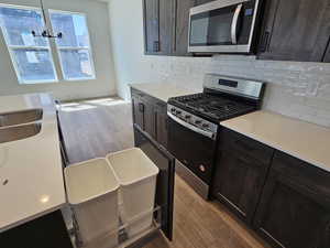 Kitchen with light wood finished floors, stainless steel appliances, hanging light fixtures, light stone counters, and dark brown cabinets