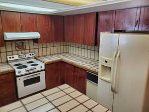 Kitchen with white appliances, tile countertops, under cabinet range hood, decorative backsplash, and tile patterned floors