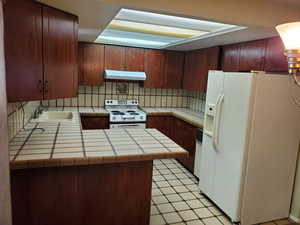 Kitchen with white appliances, light tile patterned flooring, tasteful backsplash, under cabinet range hood, and tile counters