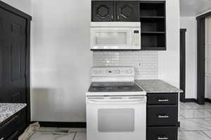 Kitchen with dark cabinets, white appliances, light marble finish floors, and light stone counters