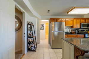 Kitchen with stainless steel appliances, dark stone counters, light tile patterned floors, brown cabinetry, and a textured ceiling