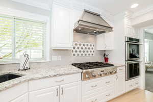 Kitchen featuring backsplash, appliances with stainless steel finishes, light stone counters, premium range hood, and ornamental molding