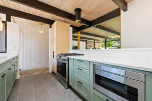 Kitchen with green cabinetry, appliances with stainless steel finishes, beam ceiling, dark tile patterned floors, and light stone counters
