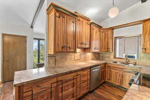 Kitchen featuring dark wood-style flooring, decorative light fixtures, brown cabinetry, stainless steel dishwasher, and vaulted ceiling