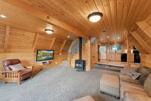 Carpeted living area with wooden walls, a wood stove, wood ceiling, stairway, and lofted ceiling