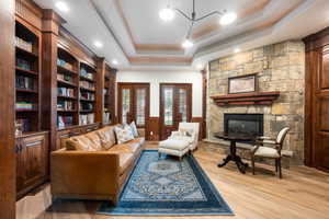 Living room featuring a stone fireplace, light wood-style floors, a wainscoted wall, a raised ceiling, and french doors