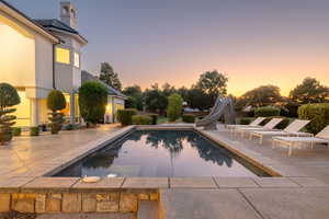 Pool at dusk with an outdoor pool, a water slide, a patio, and an in-ground hot tub