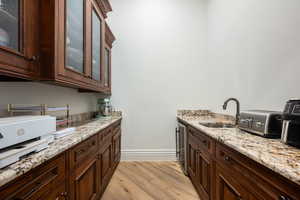 Bar area featuring light wood finished floors, light stone countertops, beverage cooler, dark brown cabinets, and glass insert cabinets
