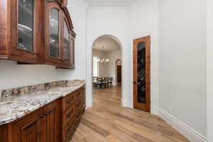 Bar with light wood-style flooring, light stone counters, arched walkways, glass insert cabinets, and ornamental molding