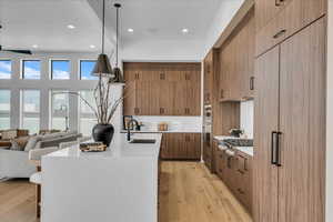 Kitchen with light stone countertops, light wood-style floors, brown cabinetry, a center island with sink, and decorative light fixtures