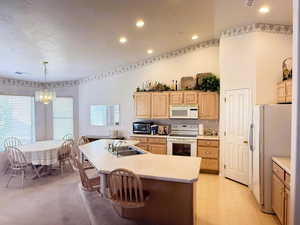 Kitchen featuring light countertops, white appliances, light brown cabinetry, a center island with sink, and recessed lighting