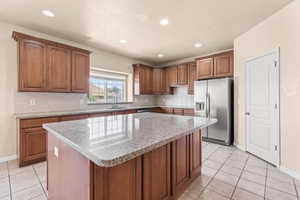 Kitchen with brown cabinetry, stainless steel fridge, light stone countertops, light tile patterned floors, and a center island