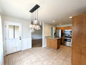 Kitchen featuring brown cabinetry, stainless steel appliances, a peninsula, pendant lighting, and a wainscoted wall