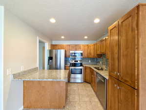 Kitchen featuring backsplash, light stone counters, stainless steel appliances, a peninsula, and brown cabinets