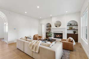 Living room featuring built in shelves, a lit fireplace, arched walkways, light wood-style floors, and recessed lighting