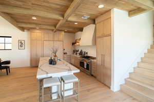 Kitchen featuring light brown cabinetry, stainless steel range, wall chimney range hood, recessed lighting, and a breakfast bar area