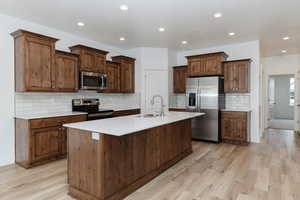 Kitchen featuring appliances with stainless steel finishes, recessed lighting, light wood finished floors, backsplash, and a center island with sink