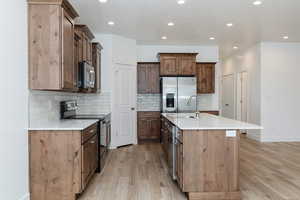 Kitchen with stainless steel appliances, light stone counters, decorative backsplash, an island with sink, and light wood finished floors