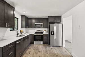 Kitchen with appliances with stainless steel finishes, light wood finished floors, dark brown cabinetry, a textured ceiling, and light stone counters