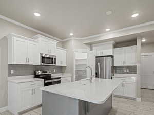 Kitchen featuring backsplash, appliances with stainless steel finishes, light stone countertops, white cabinetry, and crown molding