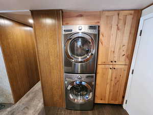 Laundry room with stacked washing machine and dryer, wooden walls, dark wood-style flooring, and cabinet space