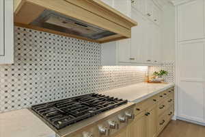 Kitchen featuring premium range hood, stainless steel gas stovetop, decorative backsplash, light stone counters, and white cabinets