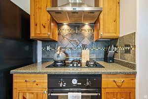 Kitchen with black appliances, wall chimney range hood, tasteful backsplash, brown cabinetry, and dark stone counters