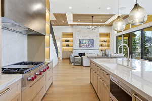 Kitchen featuring light stone counters, exhaust hood, stainless steel appliances, light brown cabinets, and hanging light fixtures