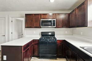 Kitchen with black gas stove, stainless steel microwave, a peninsula, a textured ceiling, and light wood-style flooring