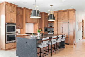 Kitchen featuring decorative backsplash, stainless steel double oven, an island with sink, hanging light fixtures, and dark stone counters