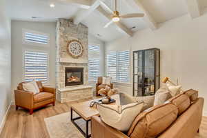 Living area with light wood-style floors, a ceiling fan, a stone fireplace, beamed ceiling, and high vaulted ceiling