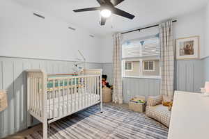 Carpeted bedroom featuring a nursery area, wainscoting, and ceiling fan